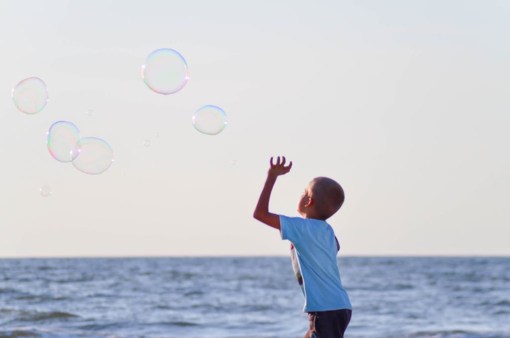 Boy in White T Shirt Playing Bubbles Near Body of Water Under Grey Sky during Daytime